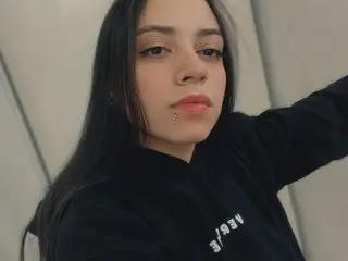 Sahara_Solis poses in a black long-sleeve top, showing her long dark hair and direct gaze against a plain grey wall.