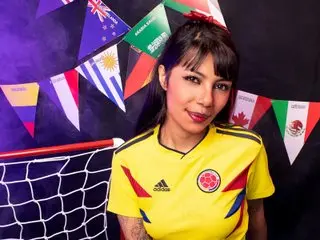 SakiAkanee wears a yellow soccer jersey and smiles in front of colorful international flags and a small soccer net.