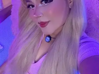 SakuraDilucas poses with her long blonde hair and heavy eye makeup, wearing a light pink top in a purple-lit setting.