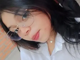 Smiling young woman with long dark hair and glasses wearing a white shirt in a bright indoor setting.