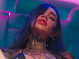 Tattooed woman in dark clothing relaxing in a neon pink and blue lit car seat with a moody atmosphere.