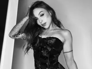 Samantaknox poses with her hand in her long dark hair, wearing a black lace corset against a plain background.