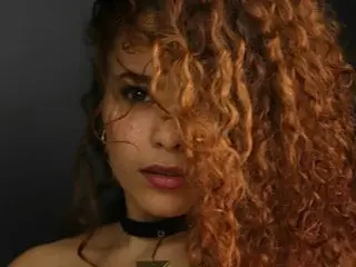 SamanthaCorreaPaz poses with her long, curly brown hair partially covering her face, wearing a black choker against a dark background.