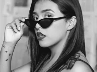 SamanthaJonhson looks at the camera with a serious expression, wearing black sunglasses and a black top against a light background.