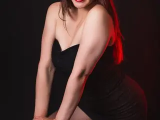 Samantha_129 poses in a black strapless dress, showing her cleavage and shoulders against a dark background with red lighting.