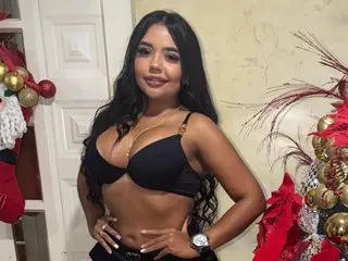 Samantha_Jhonss poses in a black bra, showing her long dark hair and cleavage against a light-colored wall and holiday decor.