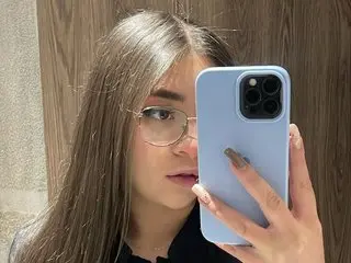 SamiiEvans poses for a close-up selfie, showing her long brown hair and glasses while holding a light blue smartphone up to her face.