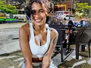 SaraHottGirl69 smiles while wearing a white tank top, showing her shoulders, arms, and cleavage in an outdoor setting.