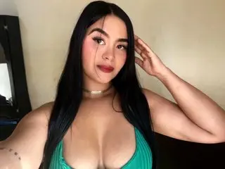 SaraLovett poses with her long black hair and red lipstick, wearing a low-cut green top that shows off her cleavage.