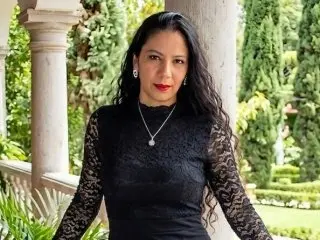 SaraSalgado poses with long dark hair, wearing a black lace long-sleeve top and a silver pendant necklace against a soft outdoor garden background.