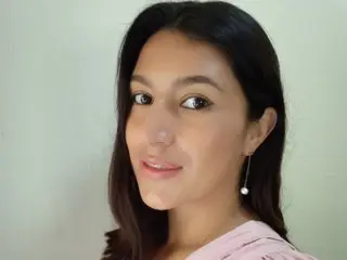 SarahFriendly is a brunette woman with long dark hair wearing a pink top and silver earrings, posing against a plain wall.