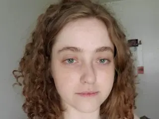 SarahSorceress looks directly at the camera with her long, curly brown hair framing her face in a close-up headshot.