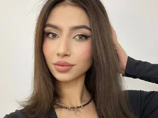 Young woman with long brown hair and glamorous makeup wearing a black top against a plain white background.