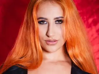 SarayMarquezz poses with long orange hair and dark eye makeup, looking directly at the camera against a red background.