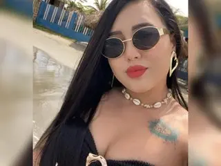 Stylish woman in black outfit and seashell necklace wearing sunglasses at a sunny beach setting.