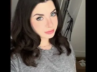 SarinaSapphire poses with a soft smile, showing her long dark hair and wearing a grey scoop-neck top.