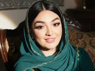 Smiling young woman in elegant green traditional hijab and gold-trimmed outfit sitting in a cozy indoor setting.