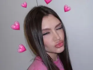 SashaNels poses with a pouty expression, showing her long brown hair and pink top against a plain white background.