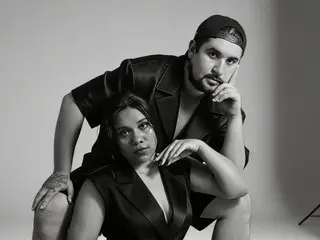 SashaSpicy poses closely with a man in black outfits against a plain studio background.