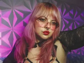 Satyndoll poses with pink hair and red lipstick, wearing a black lace top and a black choker against a purple geometric wall.