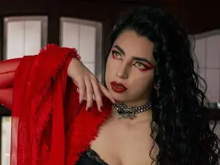 ScarlattaaDesire poses with her hands near her face, wearing a red top and a black choker with long, dark wavy hair.