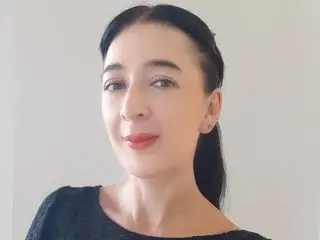 Scarlett_Blake is a brunette woman with dark hair pulled back, looking at the camera in a black top against a white wall.