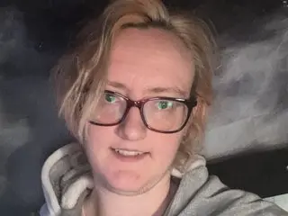 SecretKim30x looks at the camera with a slight smile, wearing a grey hooded sweatshirt and glasses, with her light brown hair swept to the side.