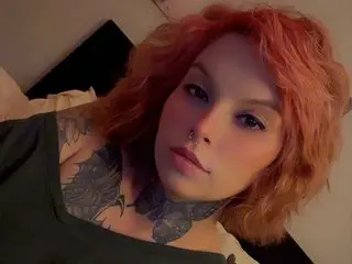 SeductionHanes poses with her wavy red hair and neck tattoos, wearing a dark green top against a plain wall.