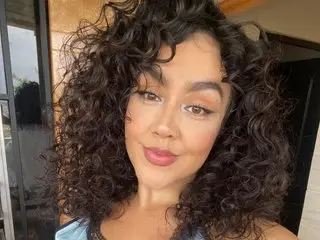 Smiling woman with voluminous curly dark hair wearing a blue top in a bright indoor setting.