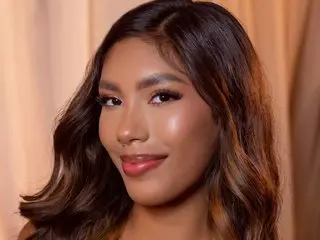 Smiling young woman with wavy brown hair and glamorous makeup posing against a neutral beige backdrop.