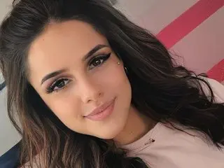 Smiling young woman with long dark hair and winged eyeliner wearing a pink top in a bright setting.