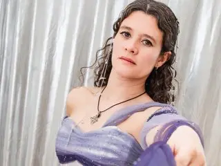 SereneLorens wears a purple strapless dress and a black necklace while posing against a silver curtain background.