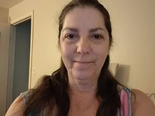 SexpertSindy looks directly at the camera with long dark hair, wearing a pink and blue striped tank top in a simple indoor setting.