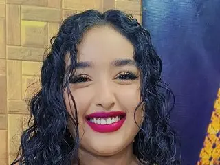 Smiling young woman with curly dark hair and bright pink lipstick posing in a warm indoor setting.