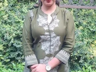SexyDayzel poses outdoors against green leaves, wearing a long-sleeved olive green tunic with silver patterns and a white neckline.
