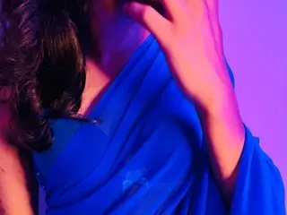 SexyOnisha poses with her hand near her face, wearing a blue wrap or cloth against a purple background.