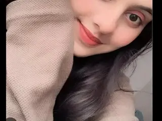Sexy_IndianSilly looks at the camera with long dark hair and a soft smile, wearing a light tan sweater.