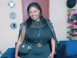 Sexykimxx smiles while wearing a black sleeveless top and black jacket, showing her hair and face in a simple indoor setting.