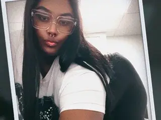 Sexylexy33 poses close-up with long dark hair and glasses, wearing a white t-shirt against a plain indoor background.