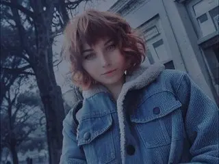 ShadowMuse poses outdoors in a blue denim jacket with a white fleece collar, looking at the camera with her wavy reddish-brown hair.