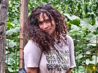 Shannom19 poses with her curly brown hair loose, wearing a light grey t-shirt against a lush green leafy background.