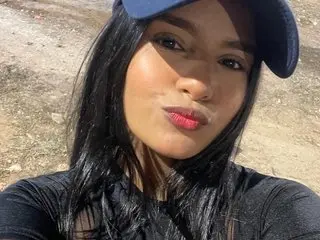Sharytt poses with long black hair and a blue cap, looking directly at the camera in a natural outdoor setting.