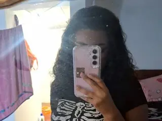 Shoriqueso poses with her long, dark curly hair, wearing a black t-shirt with a white graphic print while holding a pink smartphone up to her face.