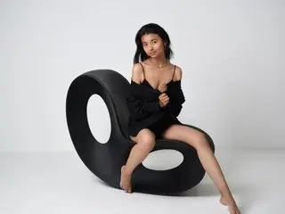 Elegant woman in a black off-the-shoulder outfit sitting on a modern sculptural chair in a minimalist studio.