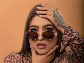 Sienna_Scott poses with her hand against her face, wearing a brown leopard print top and sunglasses with her long dark hair down.
