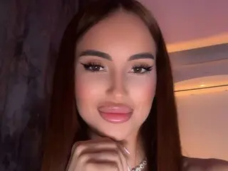 SiennaaStar poses with a hand under her chin, showing her long brown hair and soft pink lipstick.