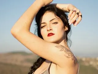 SierraSimone poses with her hands in her dark hair, showing her bare shoulders and chest against a clear blue sky.