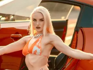 SilverCouture poses in a bright orange bikini, showing her blonde hair and toned body while standing next to a car.