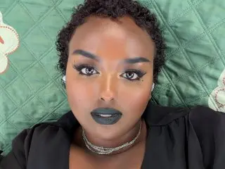 SiminFox18 looks directly at the camera with dark eyes and black lipstick, wearing a black top against a green quilted background.