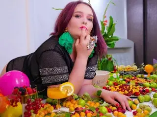 SinFulTitsDD with red hair poses behind a table of fruit, wearing a black mesh top and large green earrings.